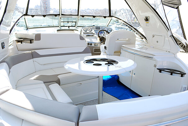 Innenansicht einer modernen Motoryacht mit einer U-förmigen Sitzgruppe in Weiß und Grau, einem runden Tisch mit Getränkehaltern und einer Steuerkonsole im Hintergrund. Die helle Ausstattung vermittelt Komfort und Eleganz.