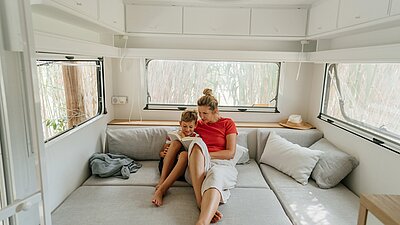 Eine Frau sitzt mit ihrem Kind auf einer grauen Couch in einem hellen Wohnmobil. Beide lesen gemeinsam in einem Buch und wirken entspannt.