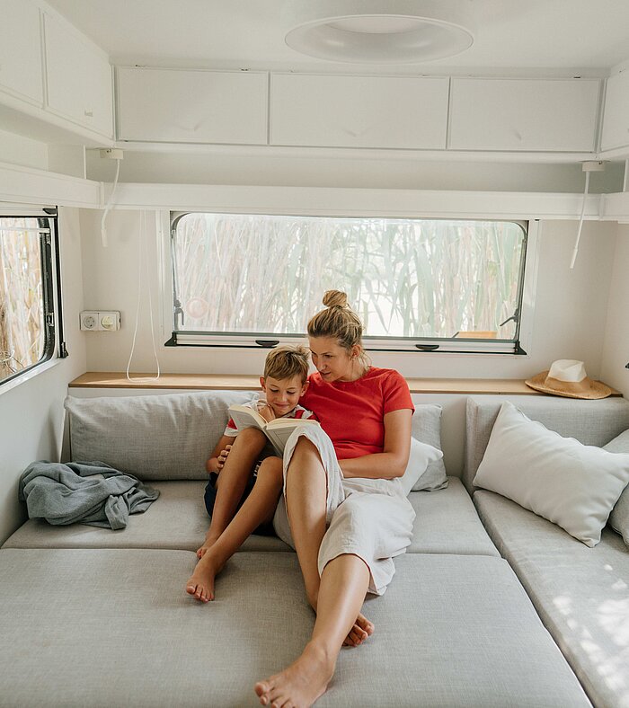 Eine Frau sitzt mit ihrem Kind auf einer grauen Couch in einem hellen Wohnmobil. Beide lesen gemeinsam in einem Buch und wirken entspannt.