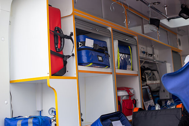 Innenansicht eines Rettungsfahrzeugs mit maßgeschneiderter Ausstattung. Mehrere farbige Notfalltaschen sind in offene Fächer und Ablagen einsortiert, ergänzt durch Schränke und Gerätehalterungen.