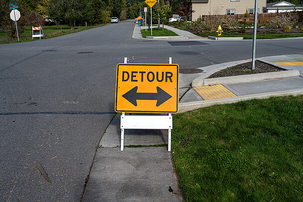 Straßenschild aus dem Kunststoffprodukt Coplast mit der Aufschrift Detour und einem Doppelpfeil nach links und rechts. Das Schild steht am Straßenrand auf einem Gehweg.