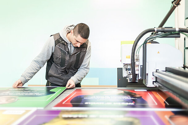 Eine Person prüft brillante Digitaldrucke auf Tafeln aus dem Kunststoffprodukt SIMOPOR. Im Vordergrund sind farbintensive Drucke zu sehen, rechts daneben die Digitaldruckmaschine.