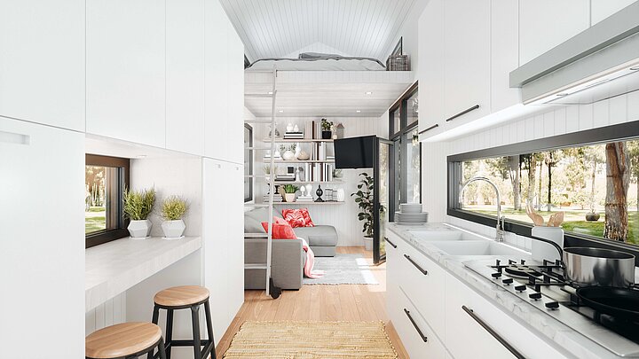 Modernes Tiny House mit heller Küchenzeile, großem Fenster und Holzfußboden. Im Hintergrund eine gemütliche Sofaecke mit roten Kissen, Regalen voller Bücher und Dekoration sowie eine Leiter zu einem Schlafbereich im Hochbett.
