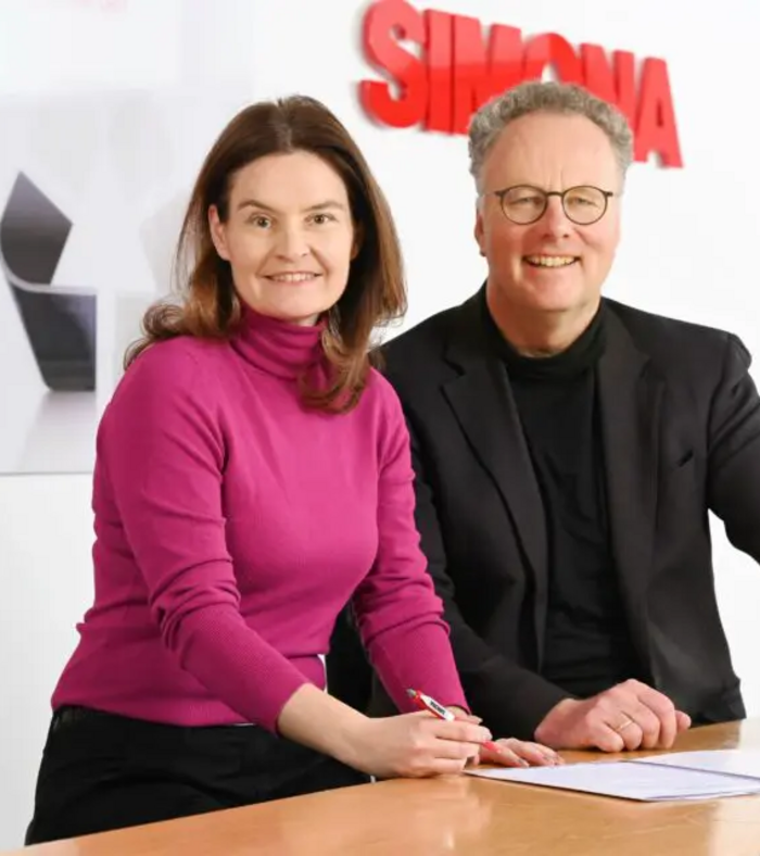Zwei Personen unterzeichnen gemeinsam Dokumente in einem Büro mit SIMONA-Logo im Hintergrund.