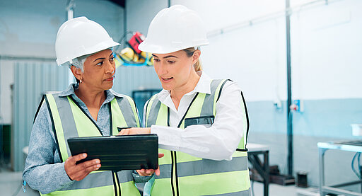 Zwei Ingenieurinnen mit Schutzhelmen und Warnwesten besprechen Produktionsdaten auf einem Tablet in einer Werkhalle.