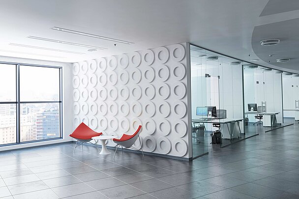 Modernes Bürointerieur mit Glaswänden und Schreibtischen mit Computern. Im Vordergrund stehen zwei rote Designstühle und ein kleiner weißer Tisch vor einer Wand mit kreisförmigem 3D-Muster.