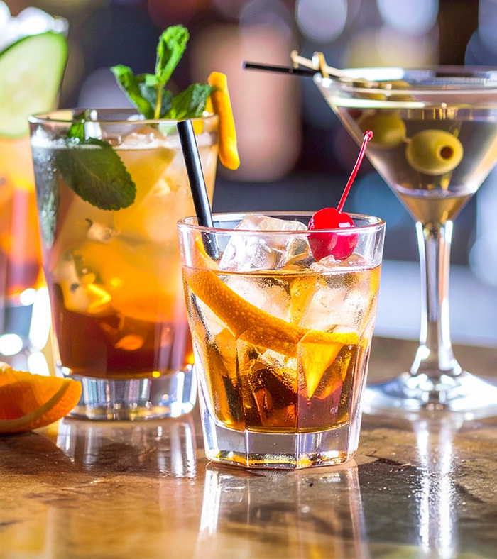 Drei Cocktails auf einer Bar: links ein Longdrink mit Minze und Gurkenscheiben, in der Mitte ein Old Fashioned mit Orangenscheibe und Cocktailkirsche, rechts ein Martini mit grünen Oliven.